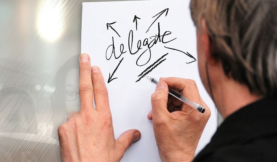 Hand writing the word delegate on a white paper with arrows pointing outward, signifying delegation