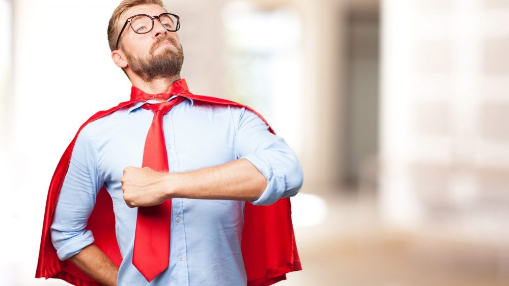 A confident man wearing glasses and a red superhero cape over a blue shirt and red tie, posing with a proud expression in an office setting