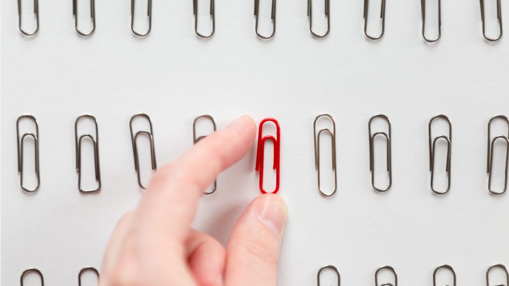 A hand picking up a red paperclip from a row of silver paperclips arranged neatly on a white surface