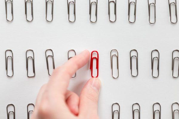 A hand picking up a red paperclip from a row of silver paperclips arranged neatly on a white surface