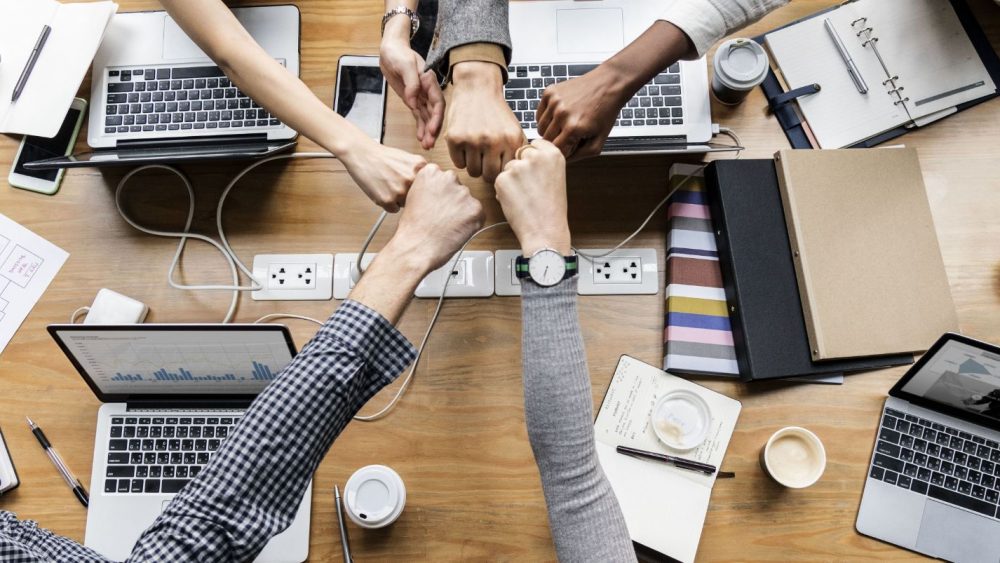 A group of people sitting around a wooden table, each with a laptop in front of them, putting their fists together in the center of the table in a gesture of teamwork